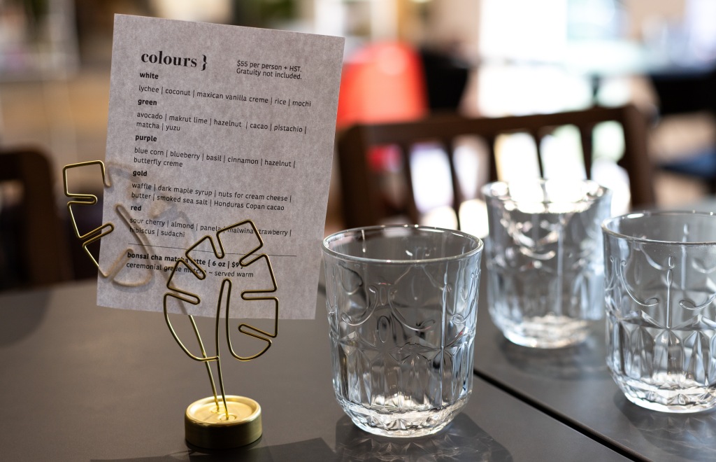 Colours menu in a leaf-shaped metal holder, on a table with three water glasses, against the blurred background of the restaurant interior. Photo by Tracy Isaacs